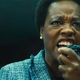 Amanda Waller, in a teal and black plaid jacket screams into a walkie-talkie.